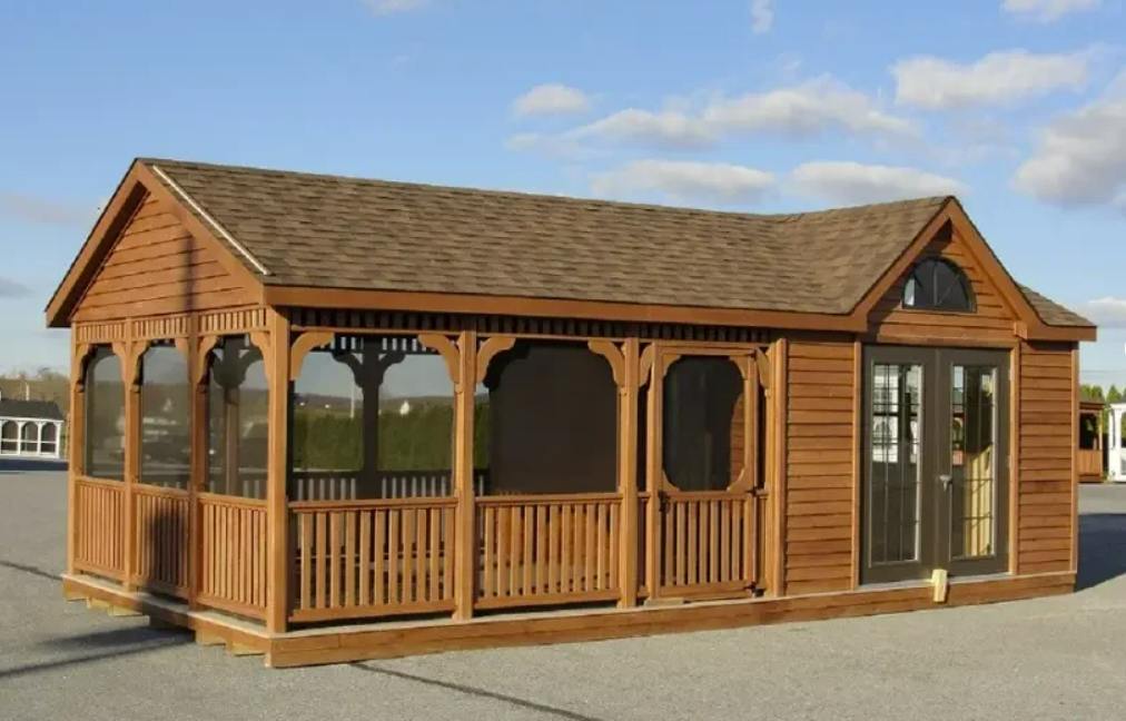 Brown wooden gazebo with screened porch, glass doors, and shingled roof under a blue sky.