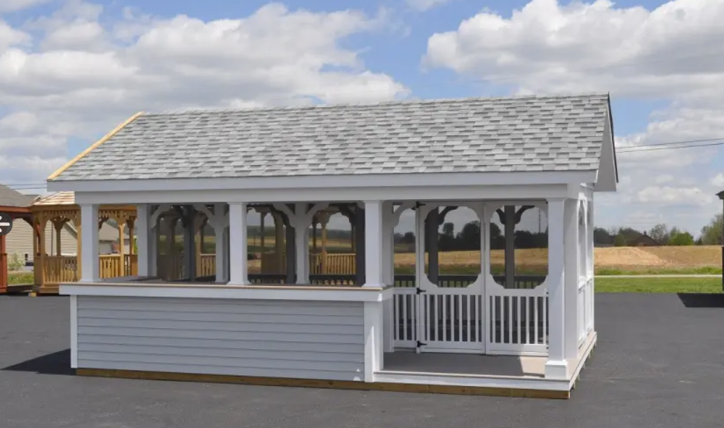 White gazebo with gray shingled roof, windows, and decorative arches on a black surface against a field under a cloudy sky.