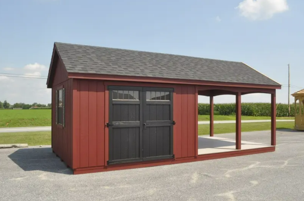 Red shed with black doors and porch. Gray roof, blue sky, and gravel ground.