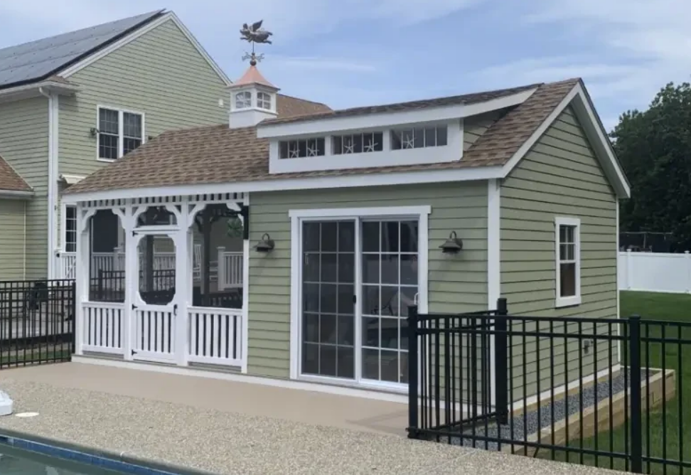 Green shed with a porch and cupola by a pool. Black fence.