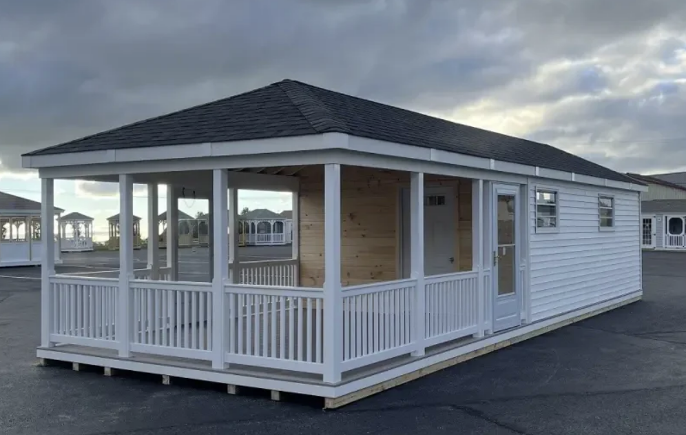 White shed with porch, black roof, and windows, set in a parking lot under a cloudy sky.