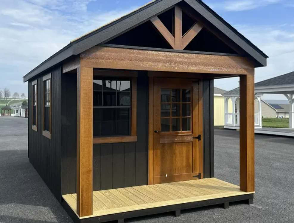 Black shed with brown trim, porch, and door. Windows on the side, set on a gravel lot.