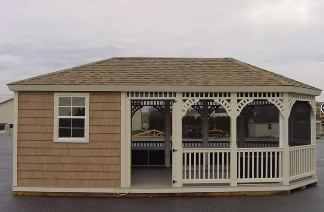 Tan wooden gazebo with a shingled roof, a window, and decorative white railings and trim.