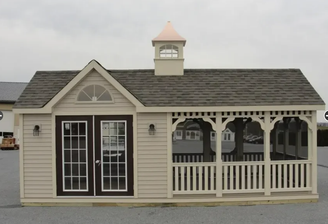 Tan shed with dark brown double doors, porch with white railing, and copper-topped cupola.