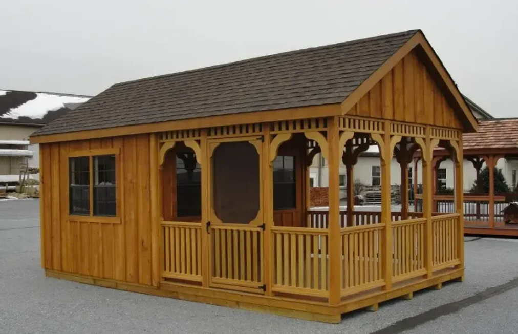 Wooden cabin with porch, brown roof, and trim. The cabin is set outside on concrete.