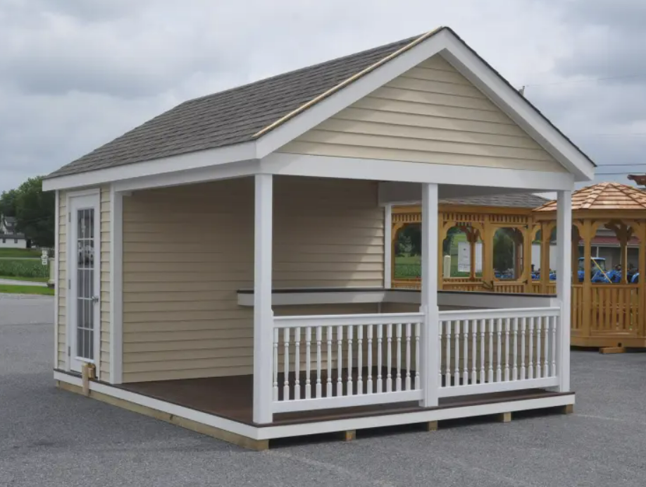 Tan shed with a porch and white railing on a gravel lot. A door is on the side, with a brown roof.