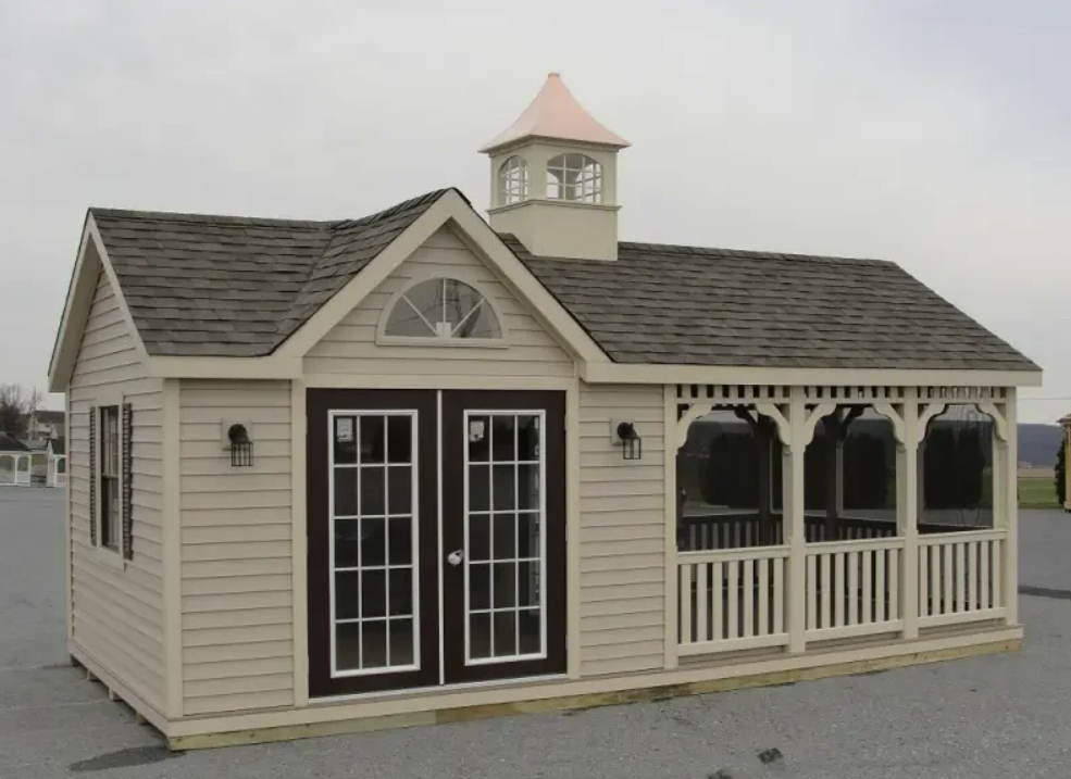 Tan shed with brown roof, double doors, porch, and cupola on a cloudy day.