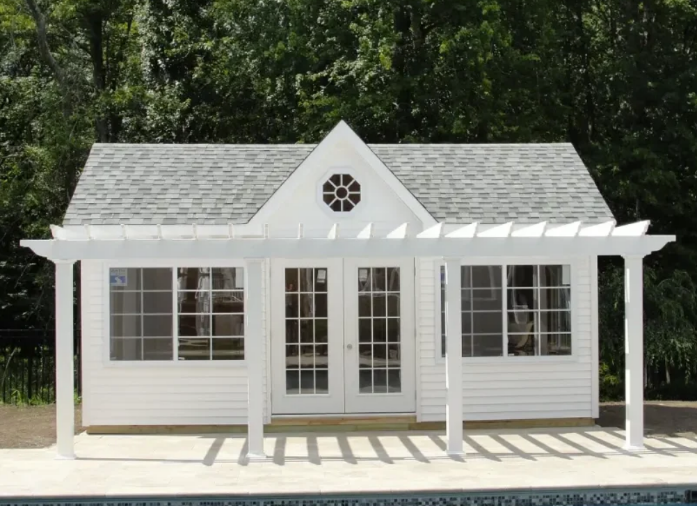 White pool house with pergola, French doors, and oval window, near a pool.