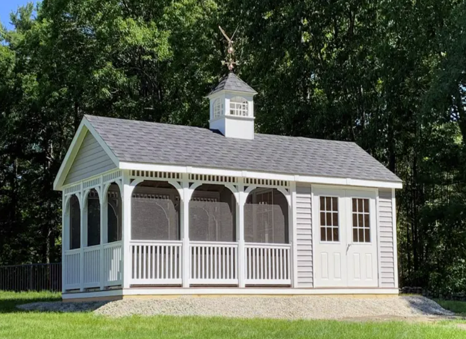 White screened-in shed with small cupola, gray roof, and arched porch. Set on a grassy lawn with trees.