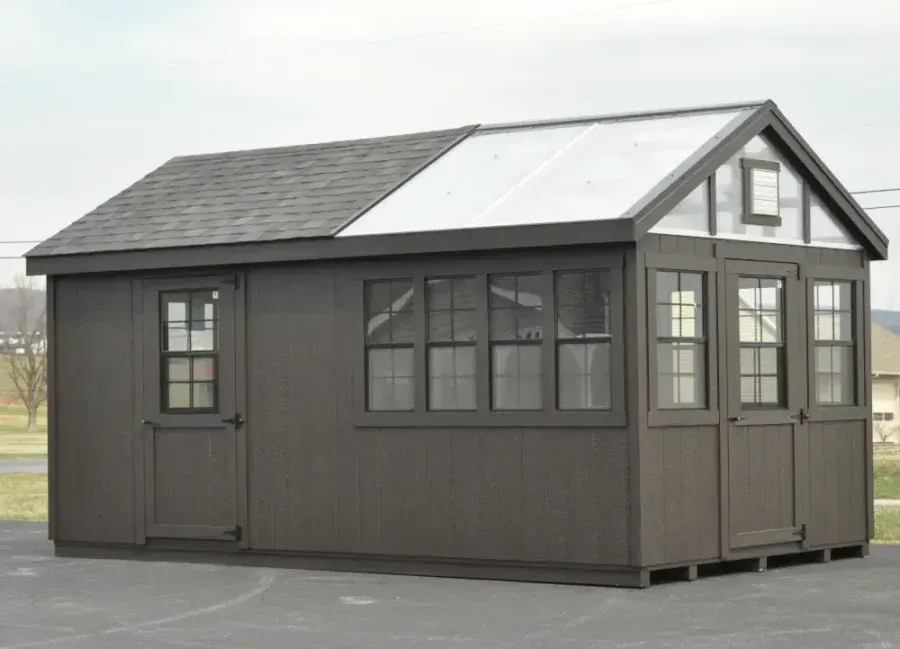 A dark brown shed with a shingled roof on the left side and a transparent, greenhouse-style roof on the right side.