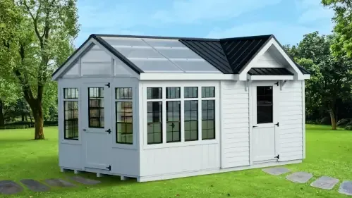 A white greenhouse-shed with large windows, a black roof, and a side door, situated on a green lawn with trees nearby.