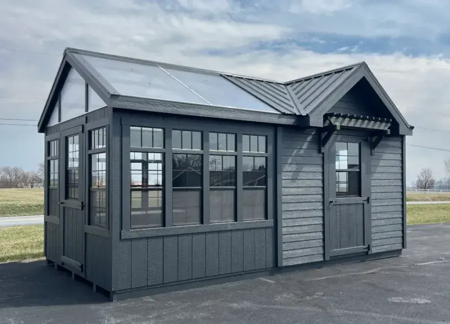 A gray, L-shaped backyard shed or greenhouse with glass roof panels, vertical siding, and a small porch over the door.