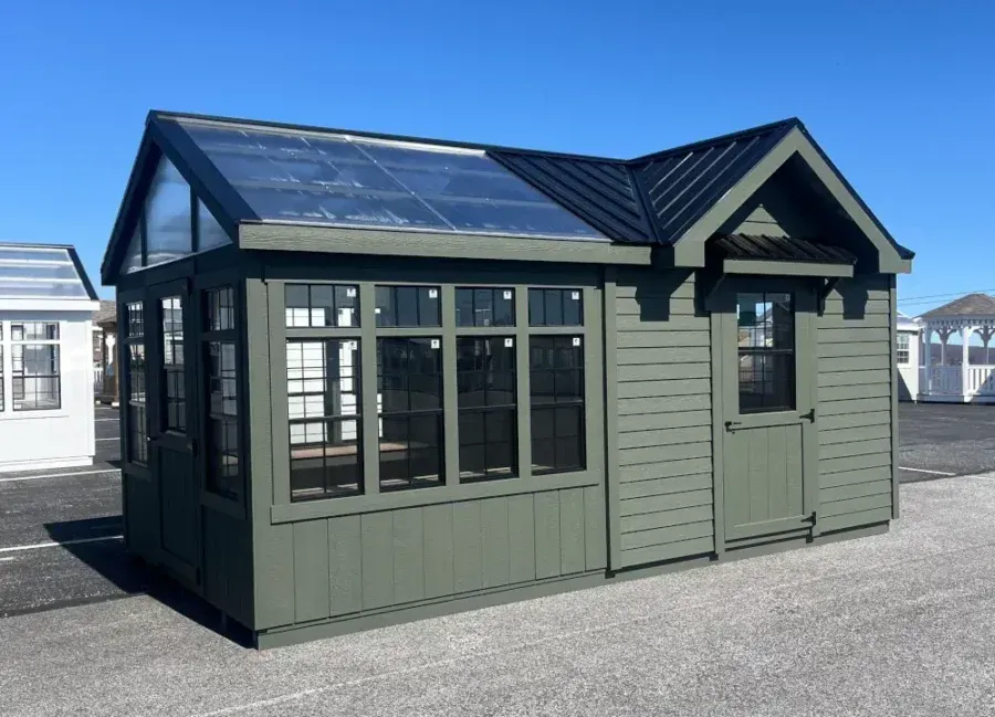 A green outdoor shed with glass panels on the slanted roof and front wall, featuring a small door, on a paved lot.