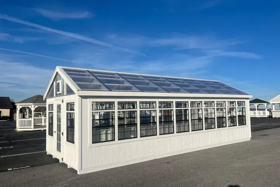 A white, rectangular greenhouse with many glass windows and a slanted roof sits on a paved lot under a blue sky.