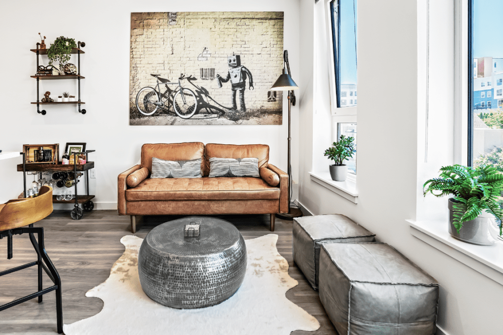 Bright modern living room with tan leather sofa, round metallic ottoman, artwork, and large windows.
