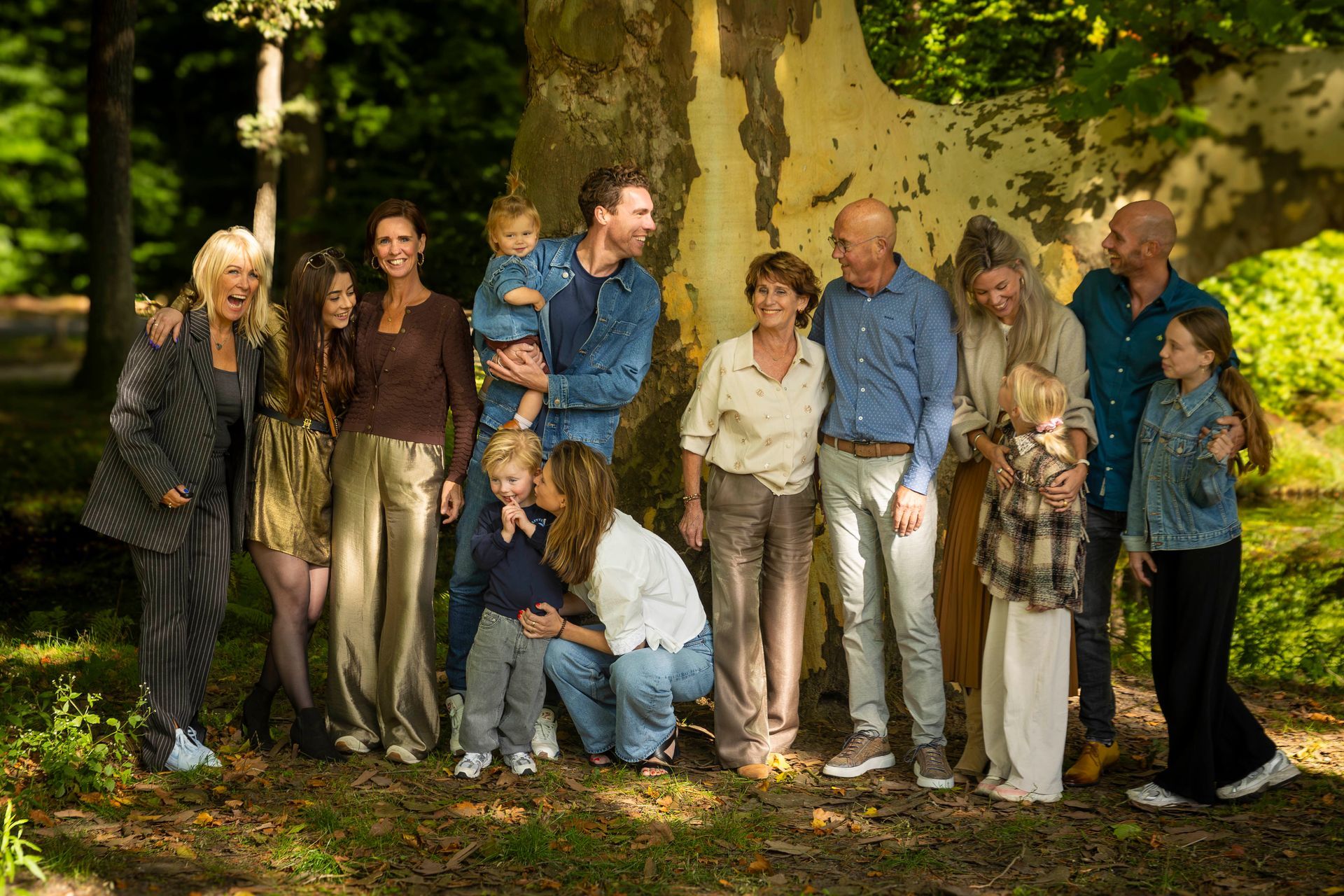 Grote familiefoto in de buitenlucht, geposeerd voor een boom. Lachend, casual gekleed, groene omgeving.