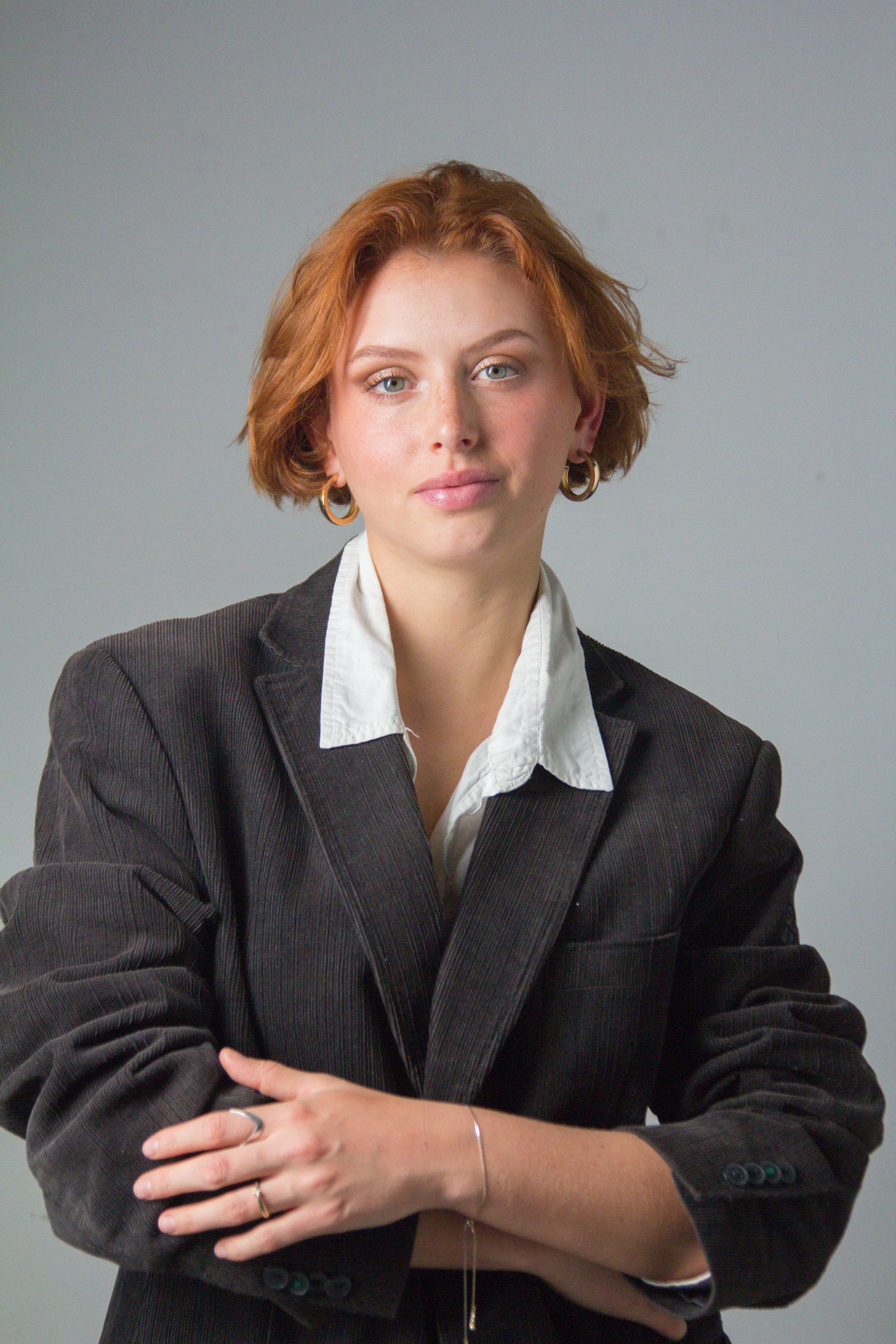 Vrouw met rood haar, gekleed in een zwarte corduroy blazer over een witte blouse, met de armen over elkaar, kijkt de kijker aan.