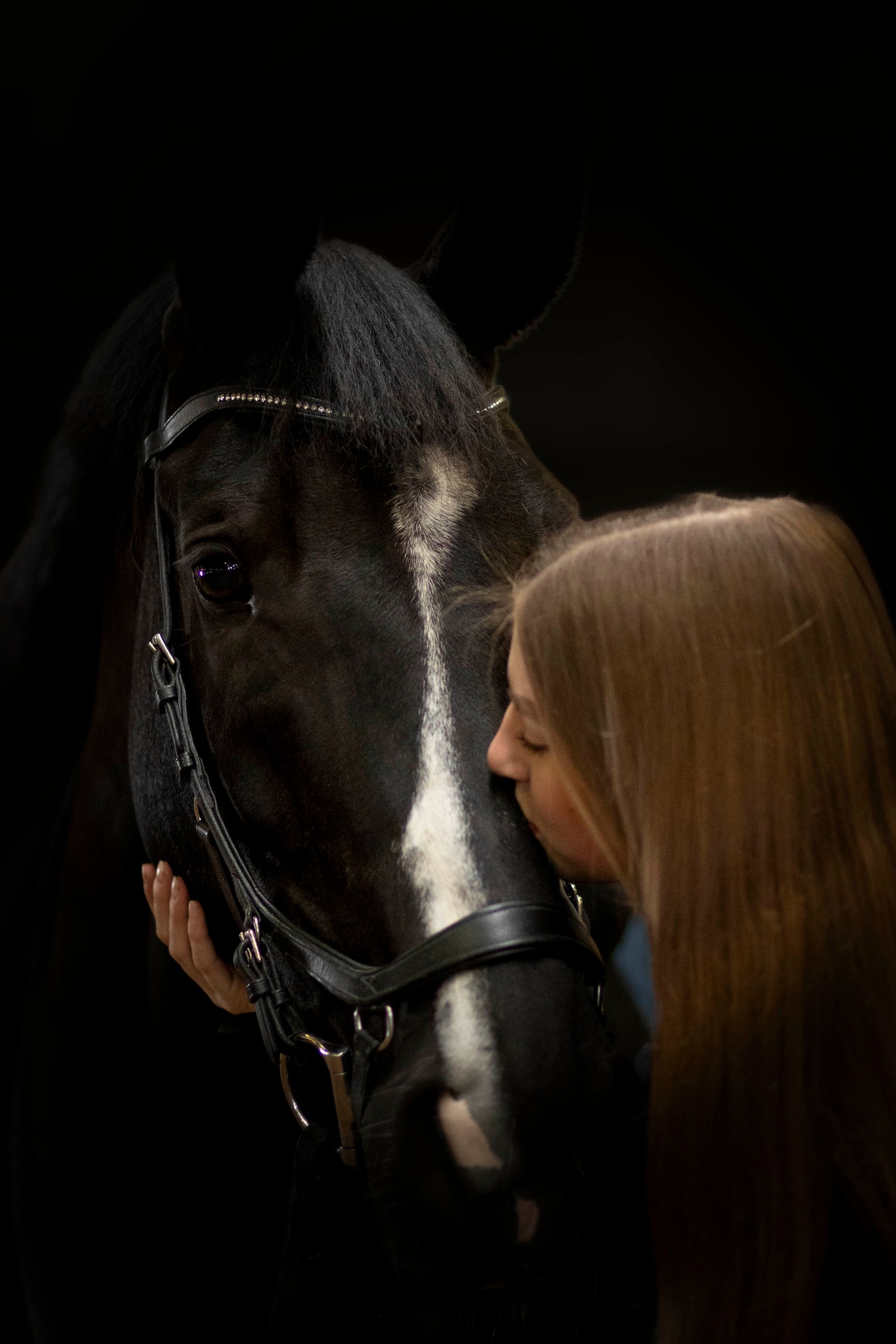 Een persoon met lang haar drukt zachtjes zijn voorhoofd tegen de witgestreepte snuit van een zwart paard in een donkere omgeving.