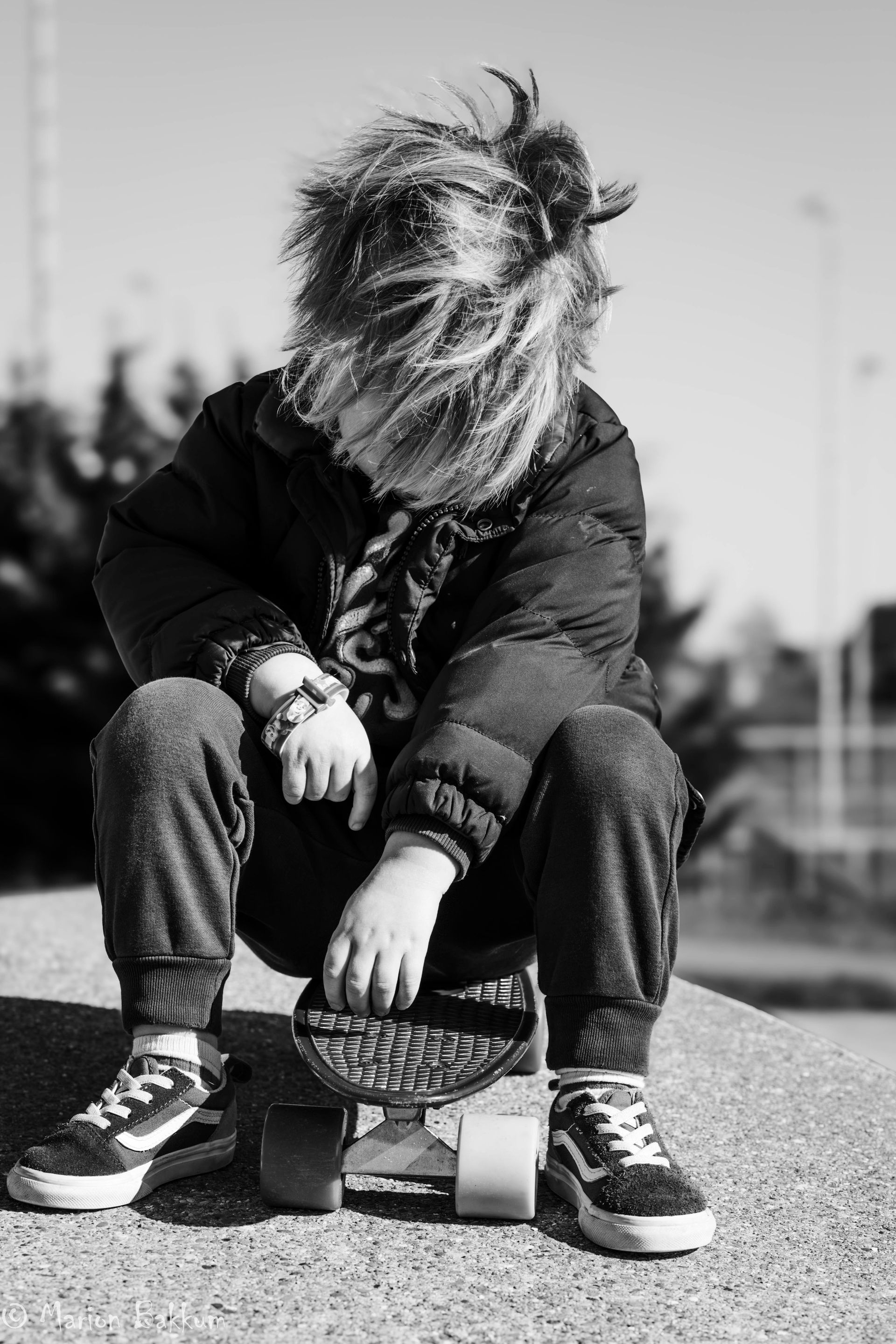 Een persoon met warrig haar zit buiten op een skateboard, gekleed in een jas en sneakers.