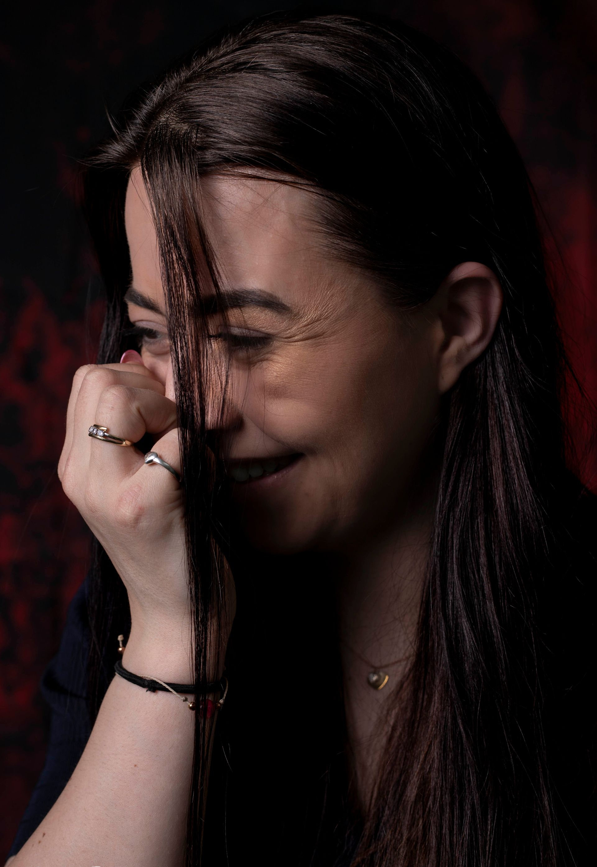 Een persoon met lang, donker haar bedekt met één hand het gezicht en draagt ​​ringen en een armband tegen een donkere achtergrond.