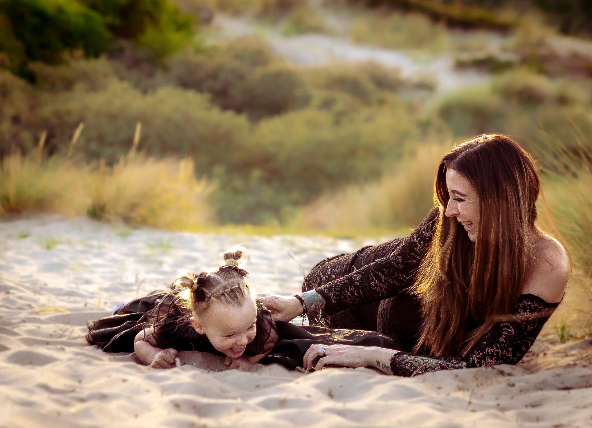 Een vrouw glimlacht naar een kind dat in het zand ligt. Beiden dragen zwarte, glinsterende jurken en zijn buiten.