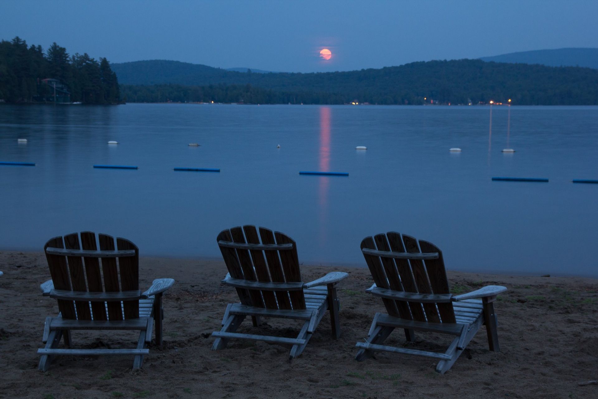 Three adirondack chairs on a beach with a full moon in the background