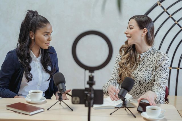 Two women are sitting at a table with microphones and a ring light.