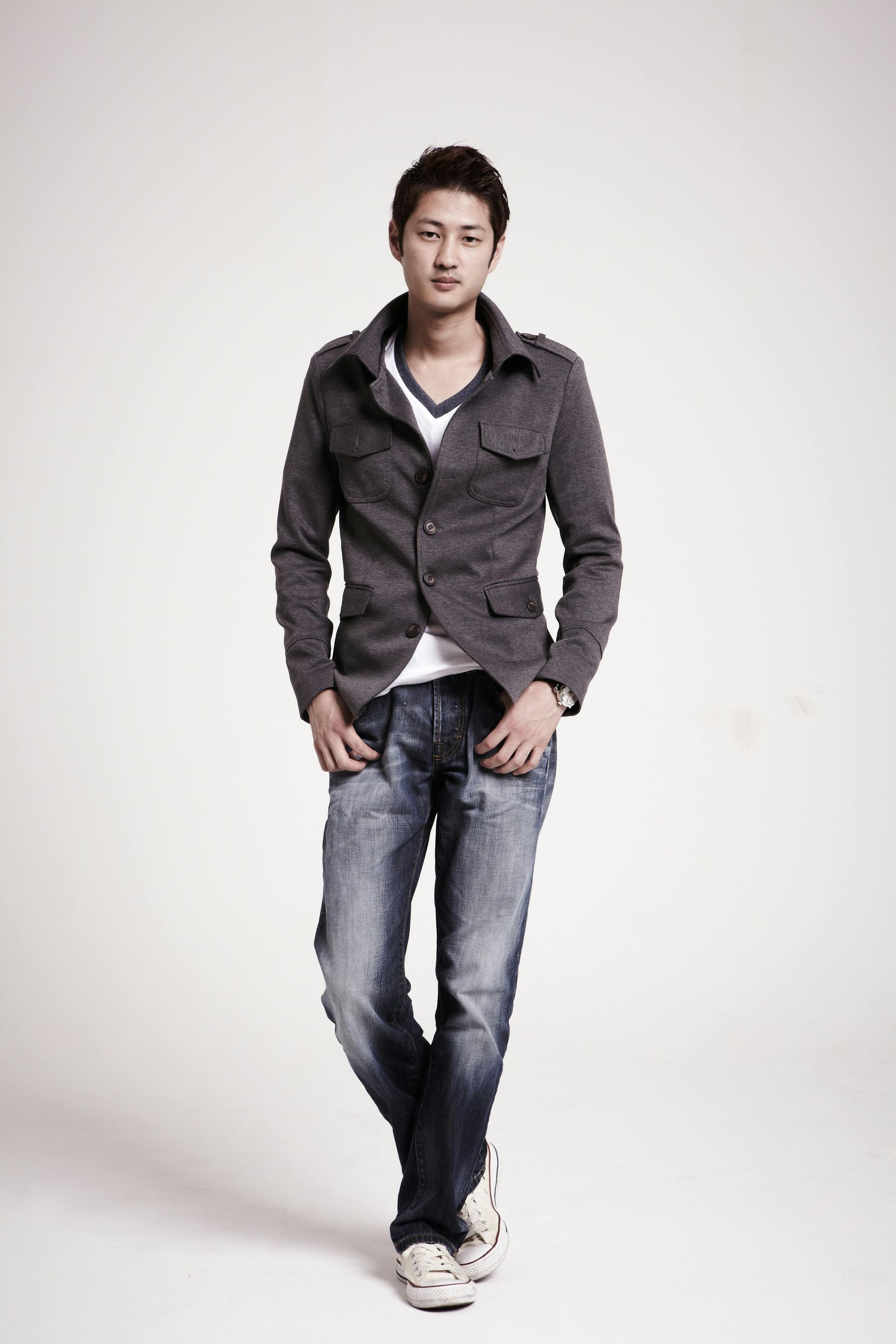 A young man stands in front of a white backdrop, wearing a gray jacket, white shirt, blue jeans, and sneakers. He has his hands in his pockets and is looking at the camera.