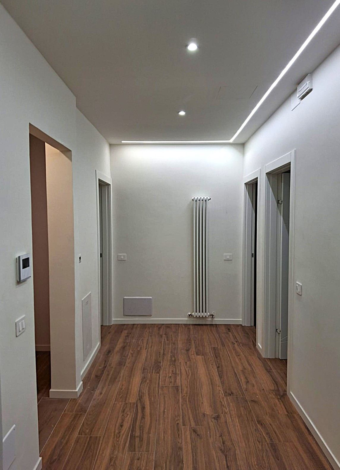 Corridoio con pavimento effetto legno, pareti bianche, faretti e porte. Un termosifone è montato a parete.