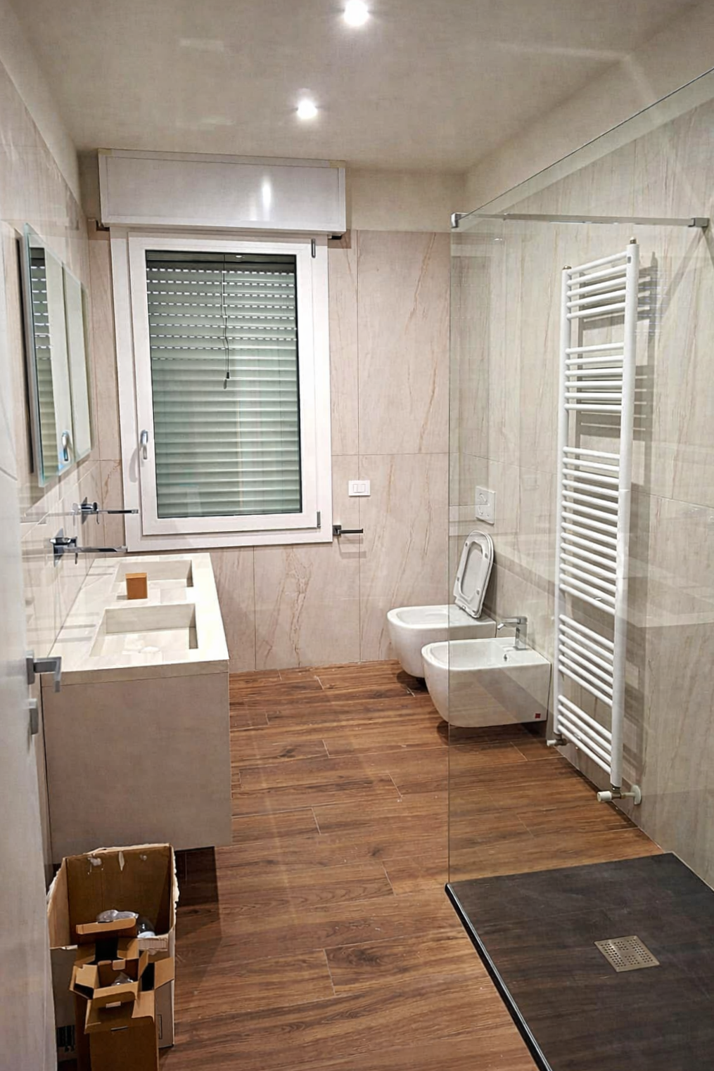 Bagno con pavimento in legno, lavabo bianco, WC e doccia. Appendiabiti appeso a uno scaldasalviette.