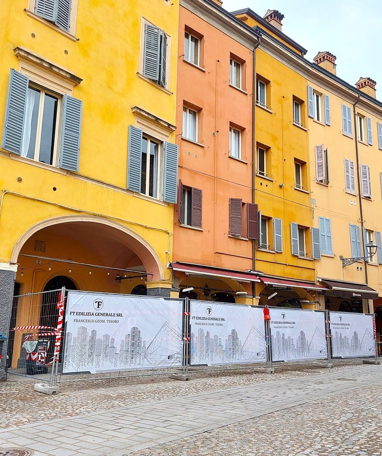 Edifici colorati lungo una strada acciottolata, con barriere di cantiere di fronte.