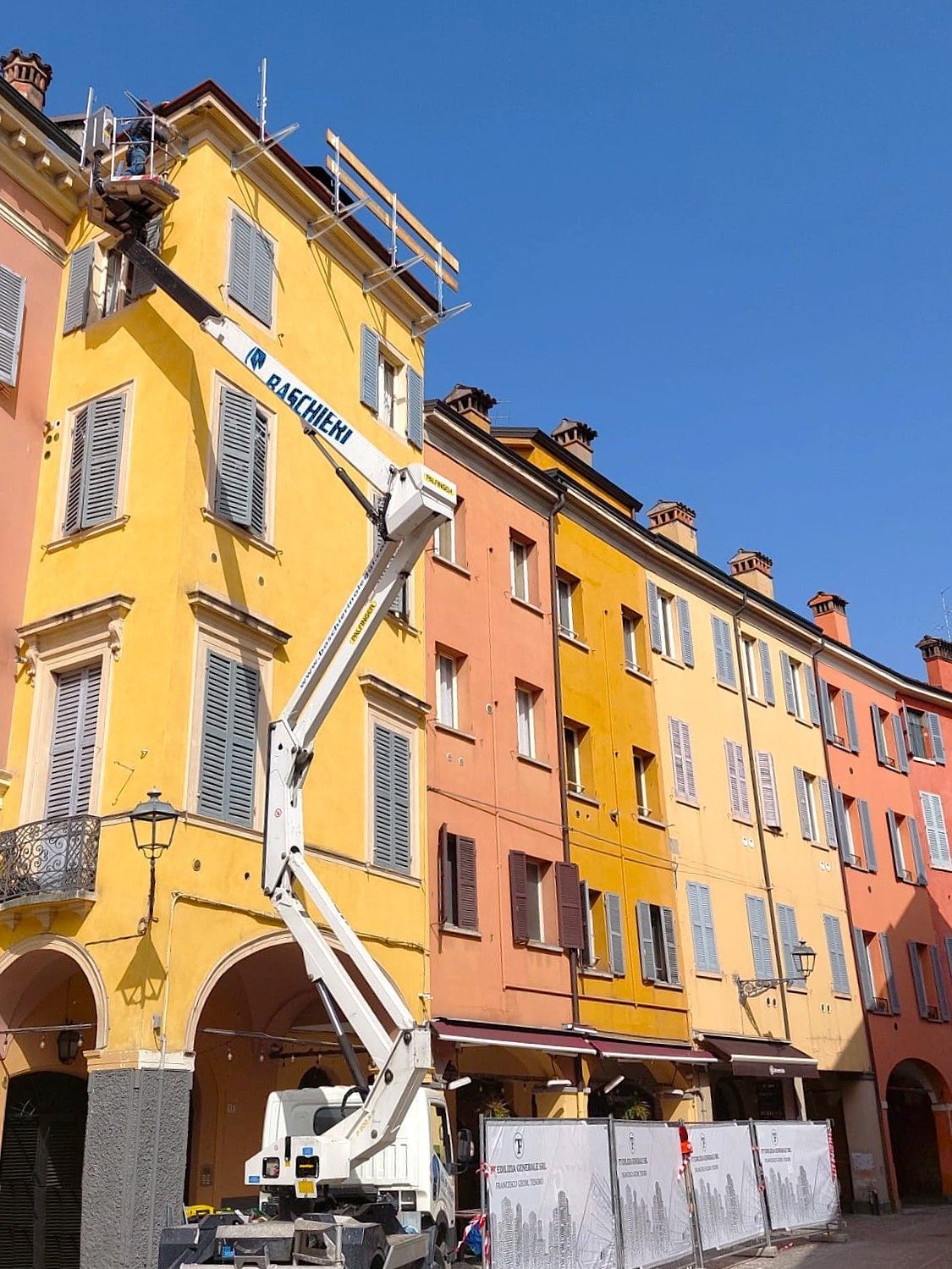 Operaio edile nel cestello dell'ascensore vicino a edifici colorati; tonalità di giallo, arancione e rosa.