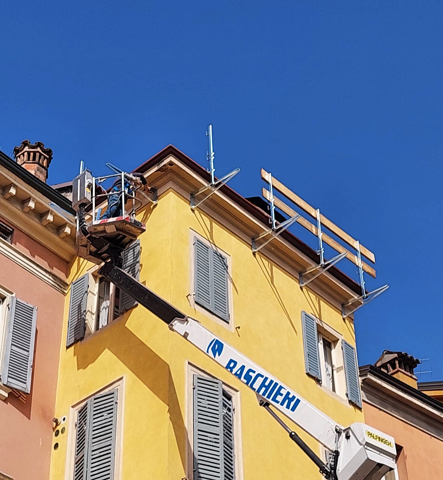 Un operaio in ascensore ripara il tetto di un edificio giallo con persiane blu, sotto un cielo azzurro e terso.