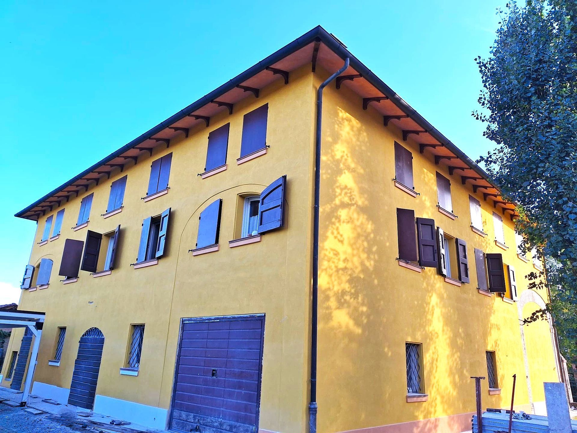 Edificio giallo a due piani con persiane e finiture marroni. Ripresa esterna soleggiata con cielo terso.