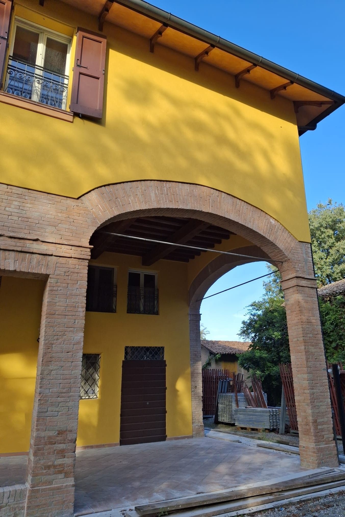 Edificio giallo con arco in mattoni e persiane marroni. Giornata di sole.