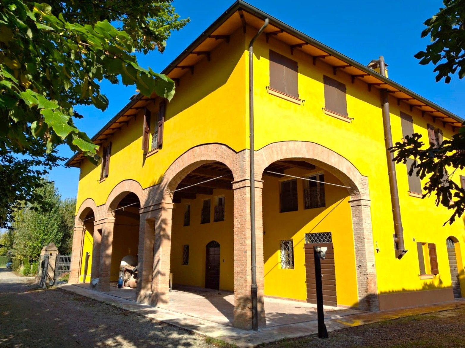 Edificio giallo con portico ad arco, persiane marroni e rami d'albero in primo piano.