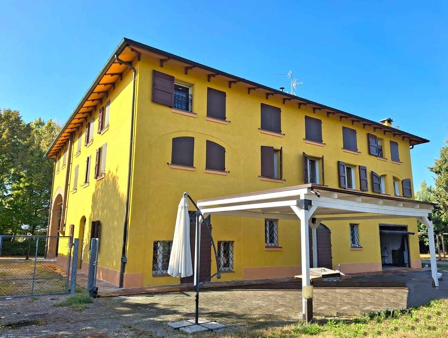 Edificio giallo a tre piani con persiane marroni e un pergolato bianco, situato in un'area aperta.