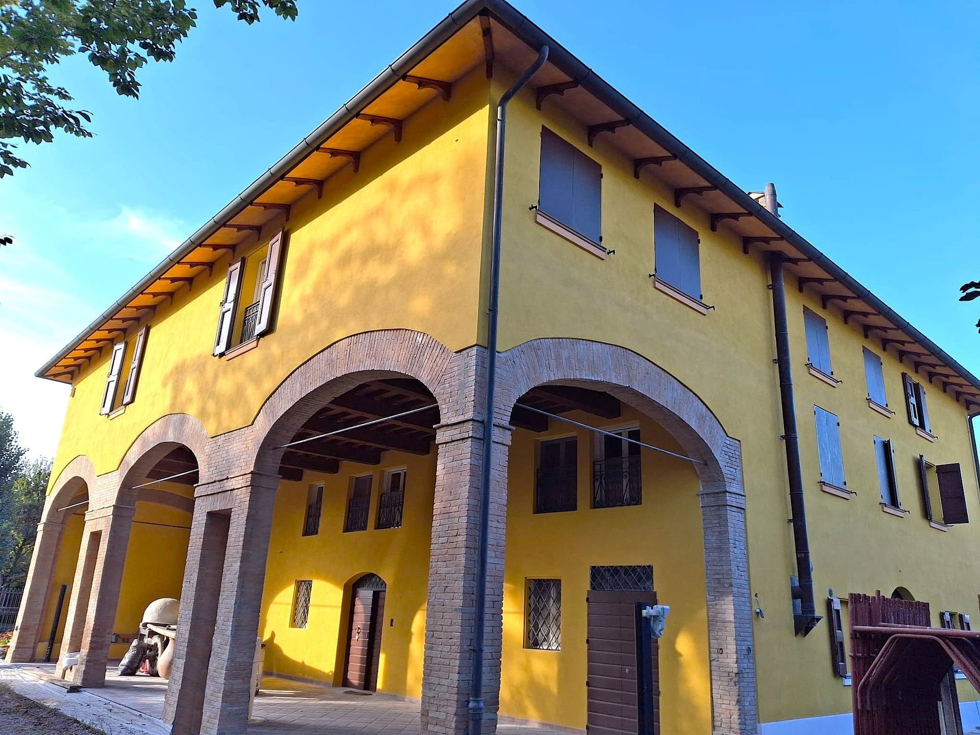 Edificio giallo a due piani con portico ad arco, persiane marroni e cielo azzurro terso.