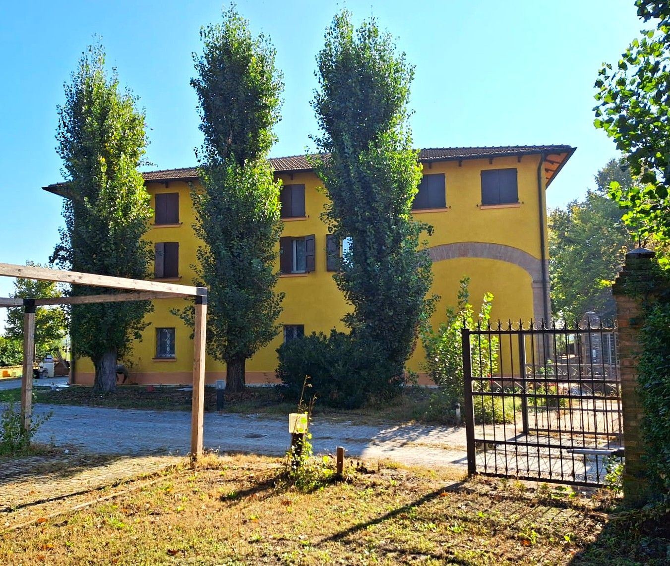 Edificio giallo con persiane marroni e alti alberi verdi di fronte, accanto a un ingresso recintato.