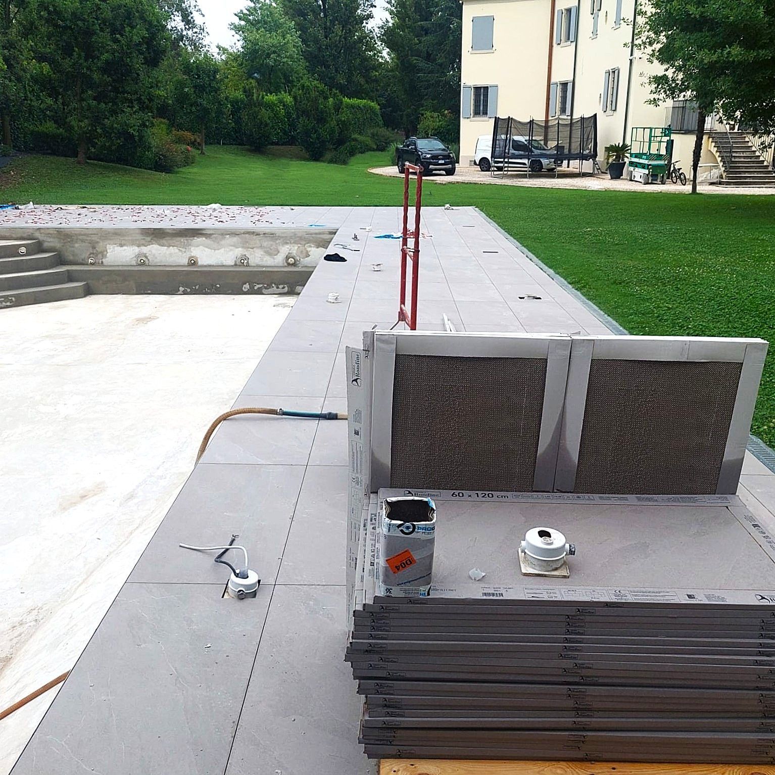 Cantiere di costruzione di una piscina con piastrelle grigie, attrezzi e un edificio sullo sfondo.