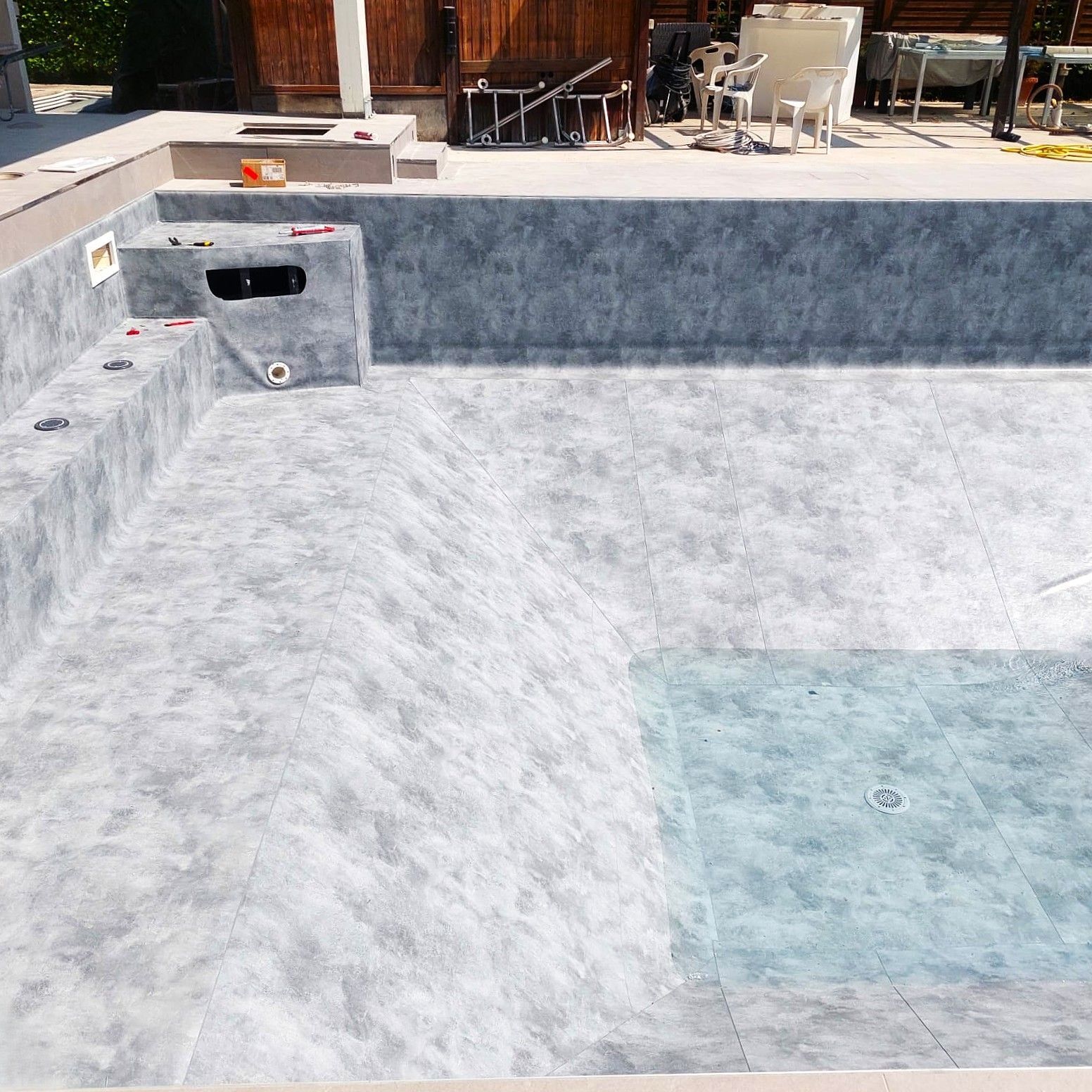 Piscina rettangolare vuota con gradini, rivestimento grigio e terrazza in cemento in un ambiente esterno soleggiato.