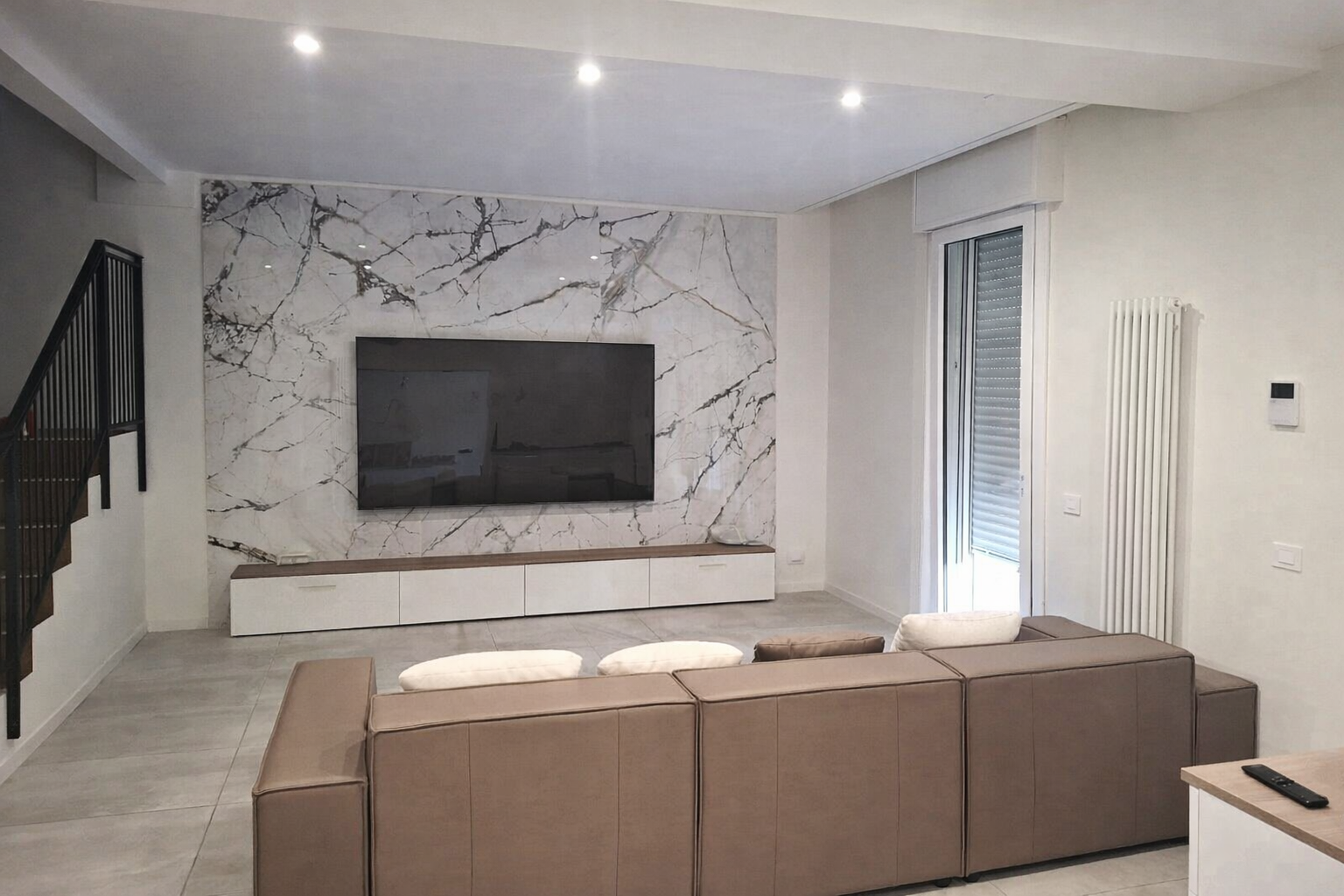 Soggiorno con ampio divano marrone, TV su parete in marmo e sedie capovolte.