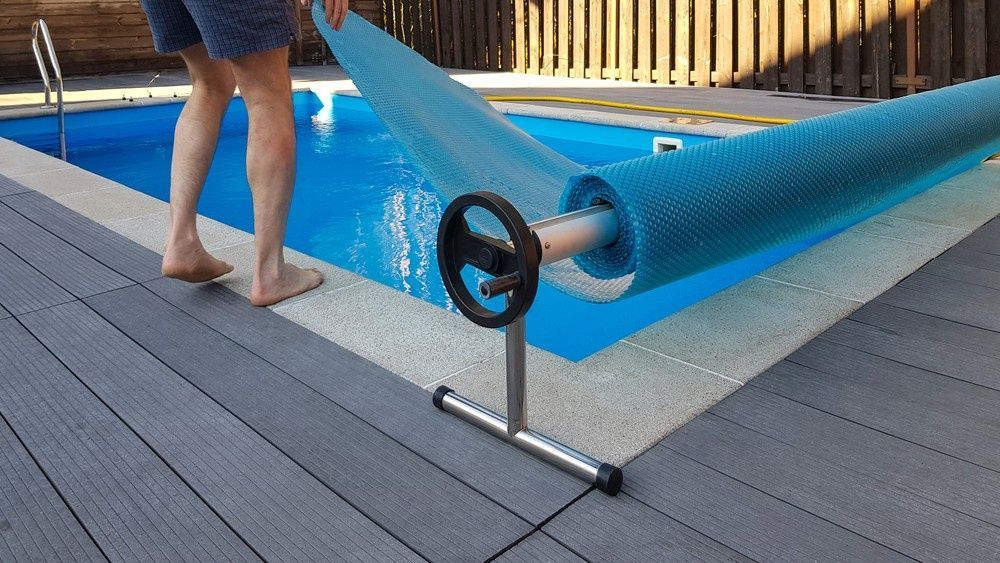 Man Unrolling a Blue Pool Cover from A Roller, Next to A Swimming Pool — Ultra Clear Pool Care in Cranbrook, QLD