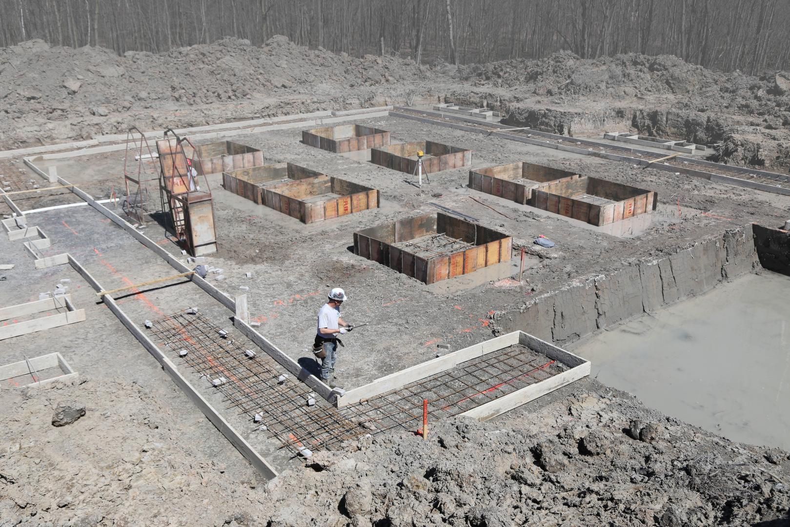 Chantier de construction avec un ouvrier debout près de coffrages en béton, de barres d'armature et de fondations partiellement coulées.