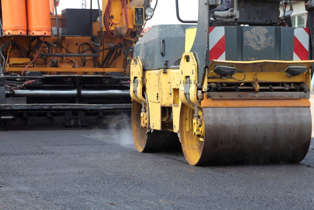 Yellow steamroller compacting asphalt road. Yellow paver in background.
