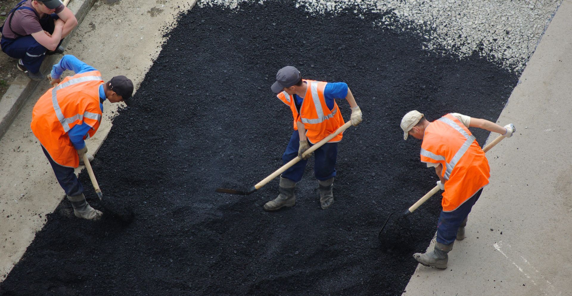 Road workers in orange vests and caps spread fresh asphalt with rakes on a street.