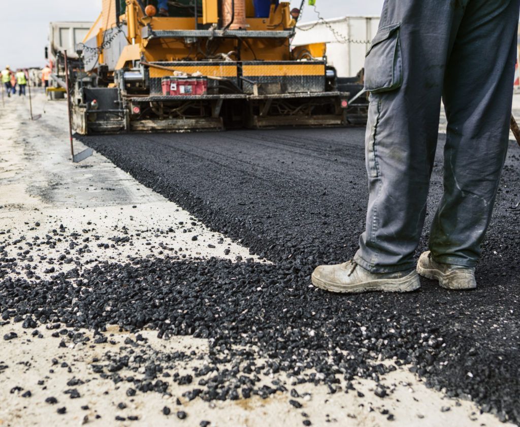 Worker standing near an asphalt paver laying black asphalt on a road.