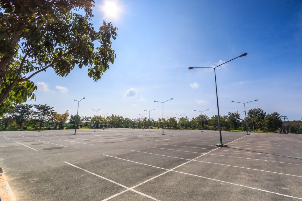 Empty asphalt parking lot under a bright blue sky with sparse trees and street lights.