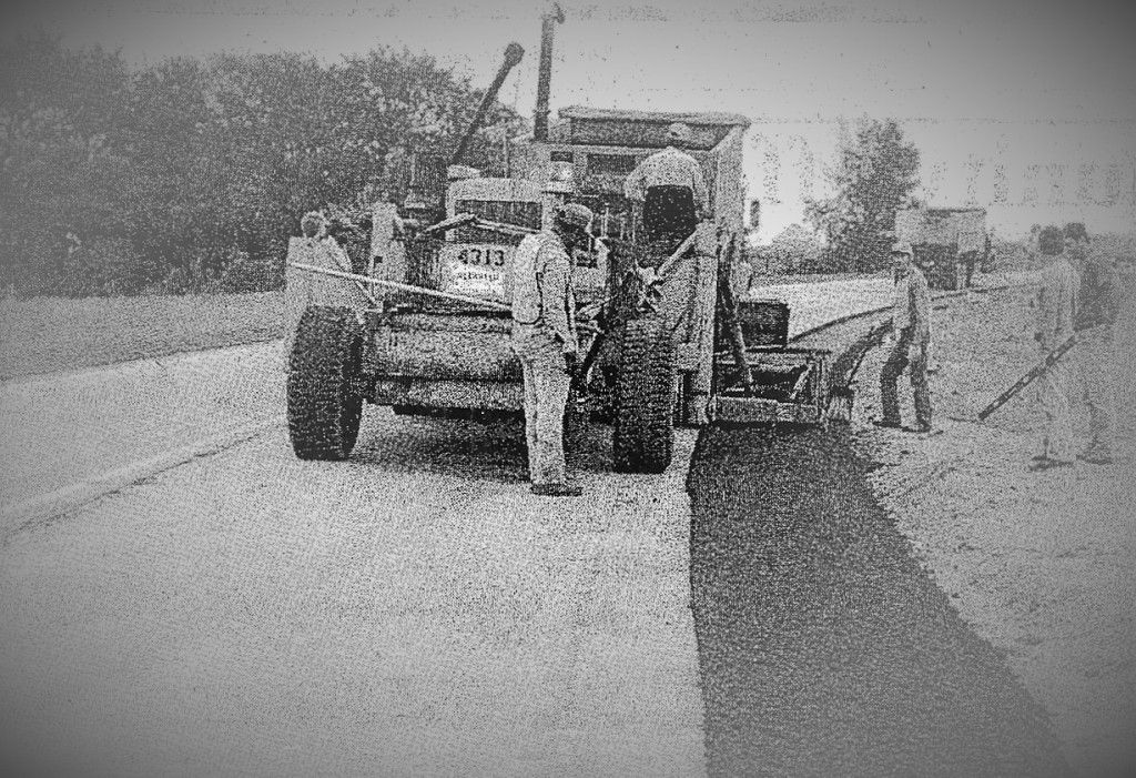 Road construction: Workers paving asphalt with a machine, alongside a rural road.