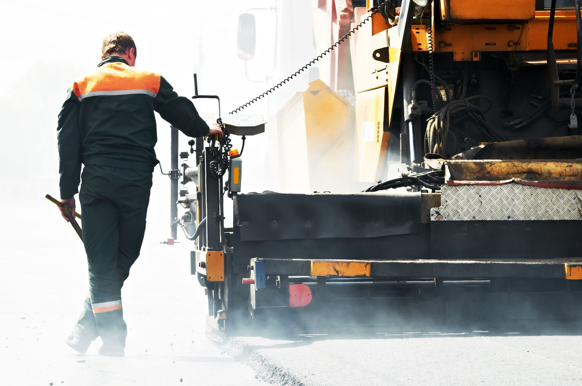 Road worker operating asphalt paving machine, misty, with orange and green workwear.